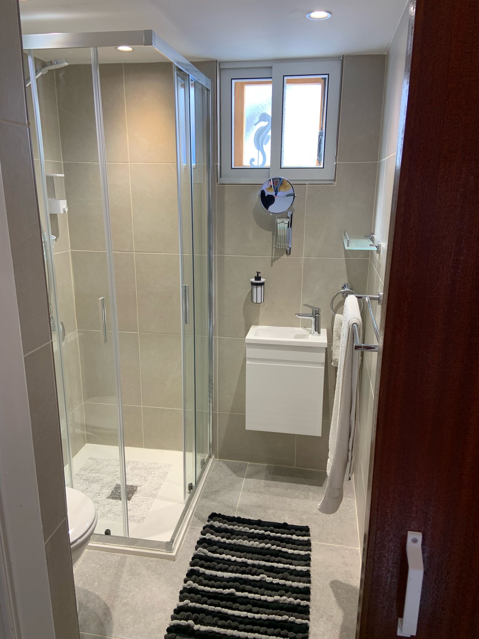 A grey tiled bathroom with a glass shower enclosure, wall-mounted basin, and window.