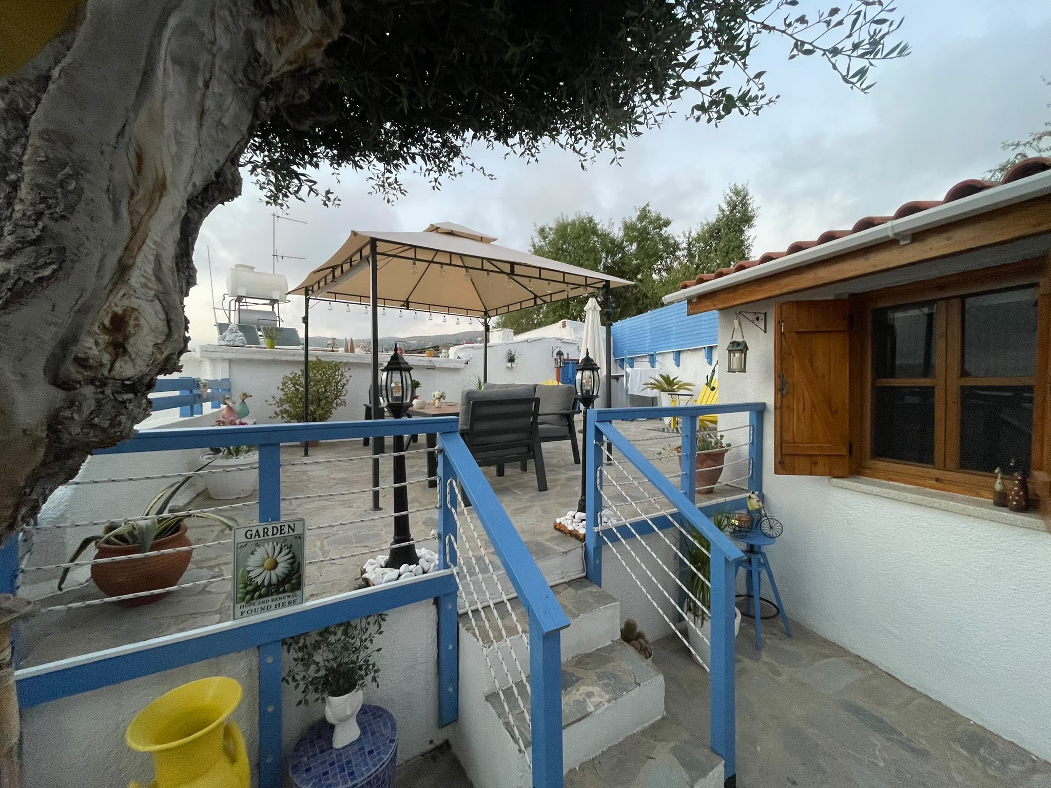 The bungalow beside a stone terrace with blue railings, a mature tree, and outdoor seating.
