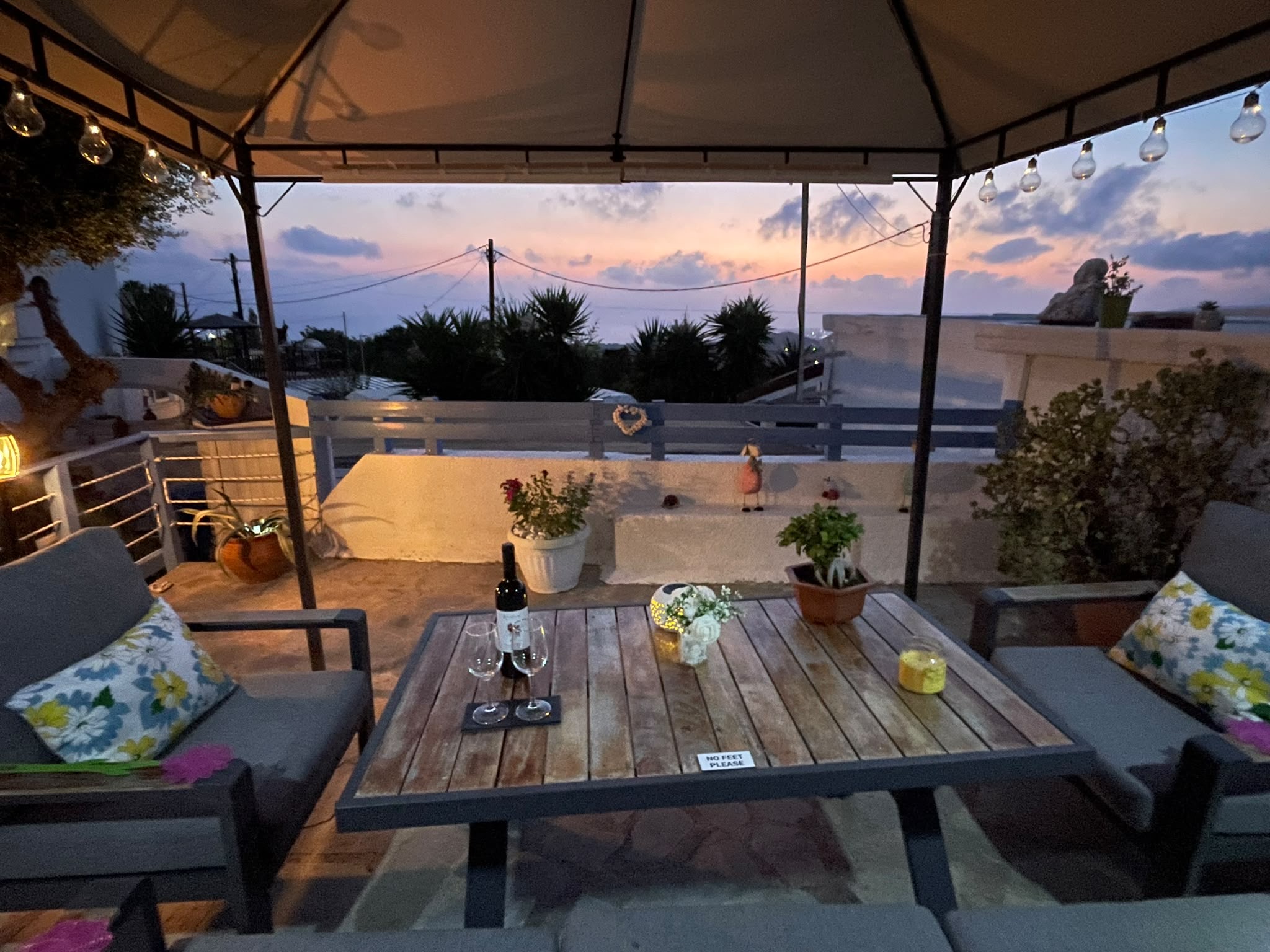 A table and cushioned chairs under the gazebo, looking toward the distant sea beneath a pink evening sky.
