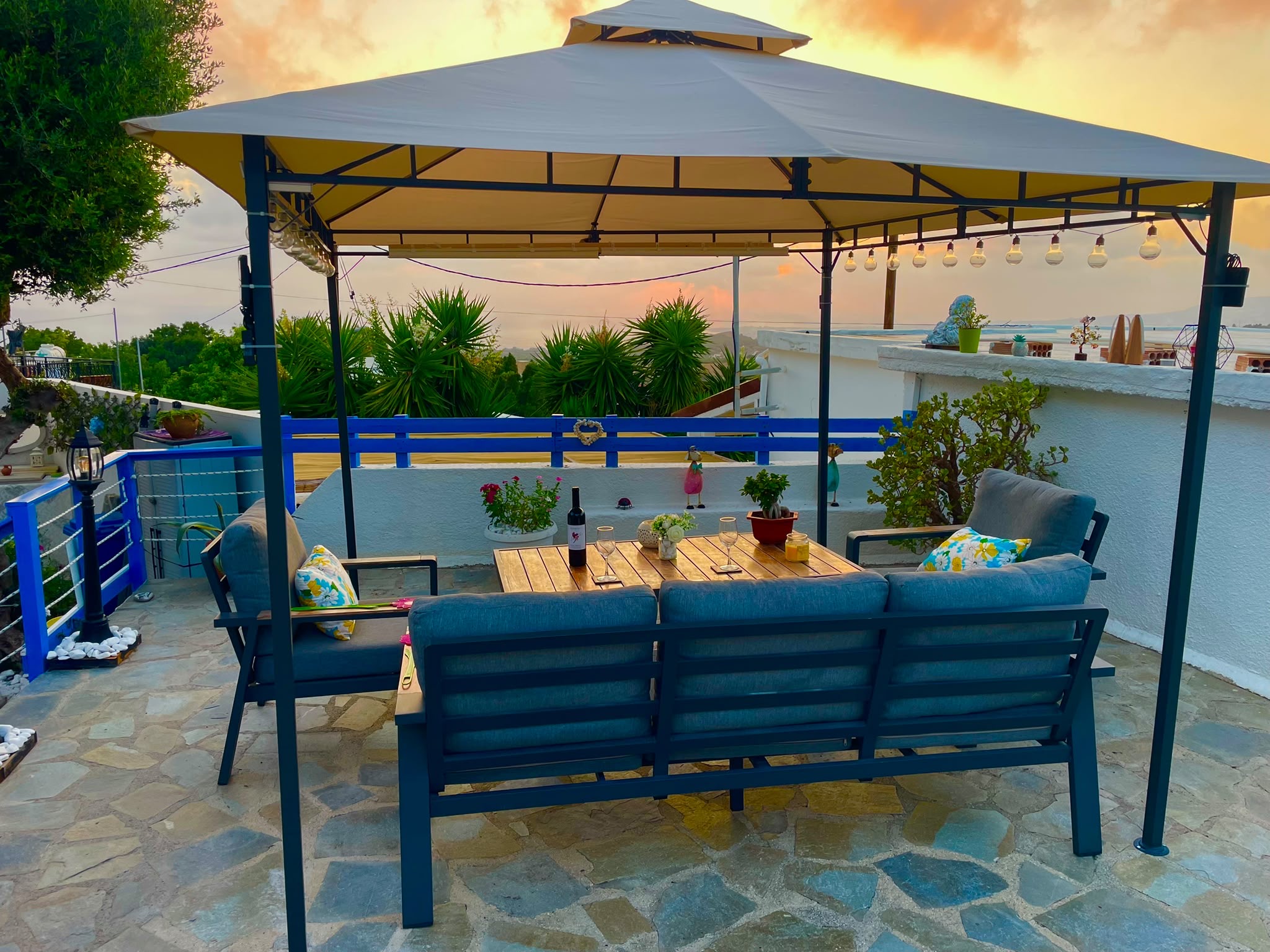 Blue outdoor seating and a wooden table beneath a gazebo on the stone terrace at sunset.